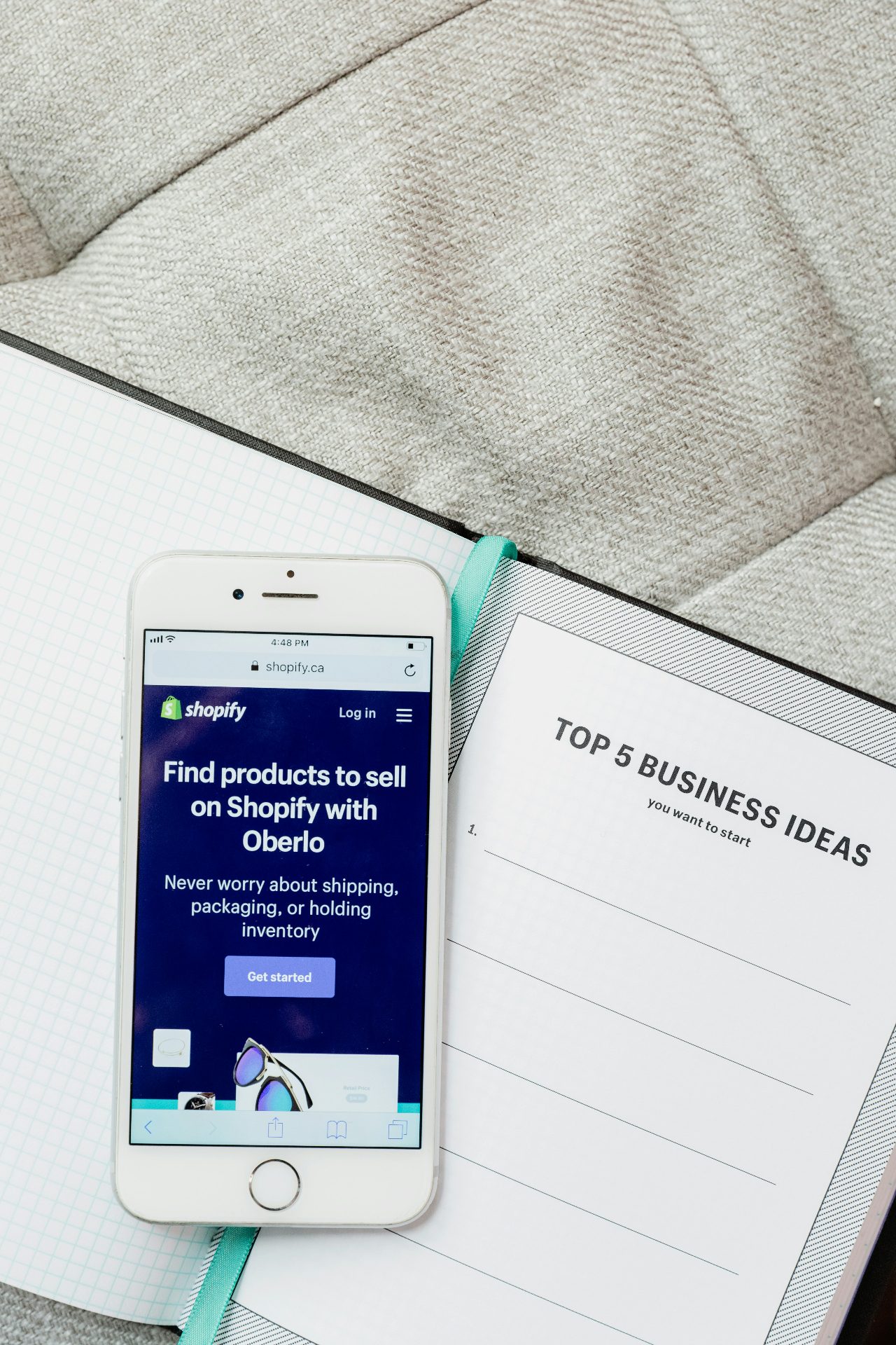 Virtual Freelance Solutions - virtualfreelancesolutions.com - How to Use AI to Write Shopify Product Descriptions in Bulk - smartphone showing Shopify app next to business ideas notebook